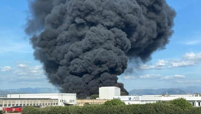 [Nessun titolo dell'immagine] - Un denso fumo nero si alza nel cielo a causa di un incendio in uno stabilimento industriale, con un'autocisterna su una strada vicina e degli edifici in primo piano.